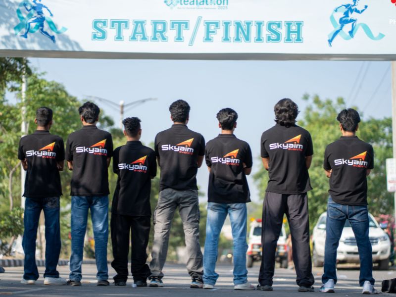 Eight people wearing black Skyaim shirts stand in a row at a start/finish line arch, facing away on a sunny street, ready to race.
