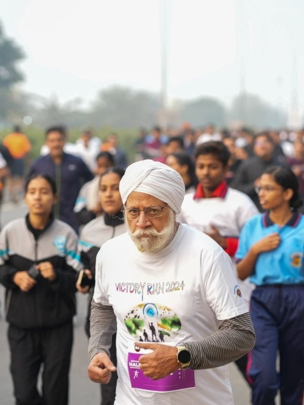 Older man in a white turban and glasses running in a crowded road race, wearing a 'Victory Run 2024' shirt and a purple bib.