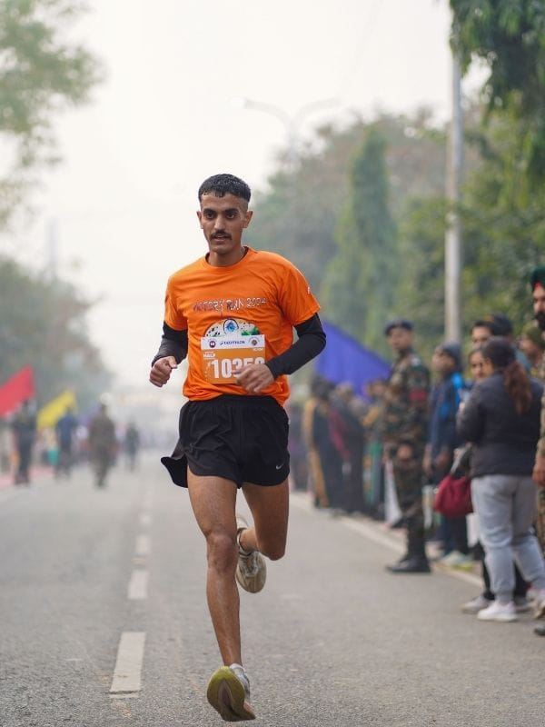 Male runner in an orange shirt and black shorts sprinting down a city street during a race, bib number 1025 visible, spectators watching nearby.