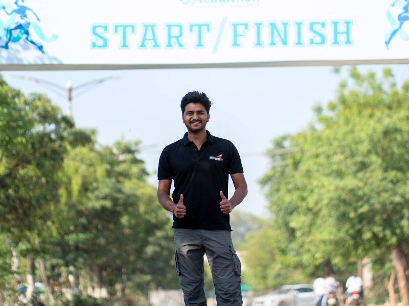 Man in a black polo and gray cargo pants stands under a bright START/FINISH banner outdoors, giving two thumbs up and smiling as trees line the street.