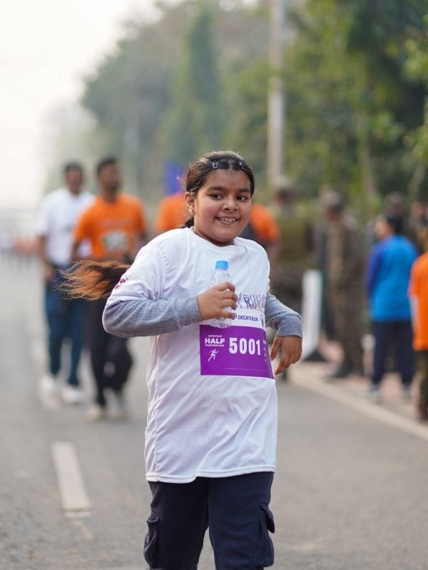 Smiling girl in a white shirt runs in a road race, bib 5001, holding a water bottle.