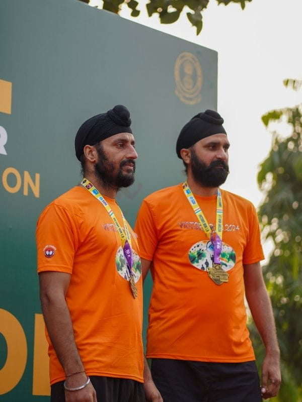Two men in orange shirts with black turbans wearing gold medals at an outdoor event.