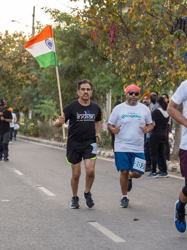 Runner carrying an Indian flag on a street race, wearing a black t-shirt and shorts, with other runners nearby.