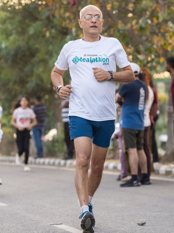 Man with glasses running on a city street wearing a white t-shirt that reads 'tealathon 2025' and blue shorts, during a race.