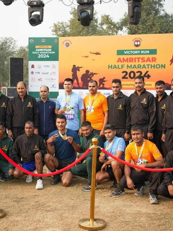 Group of runners and organizers posing with medals in front of the Amritsar Half Marathon 2024 banner, with a red rope barrier and event signage nearby.