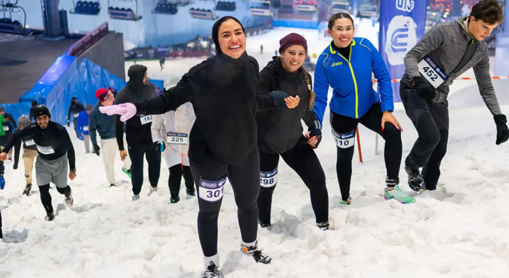Group of smiling runners with bib numbers climb a snowy slope during a winter race, cheering as they ascend with a blue-tinted venue in the background.