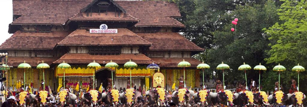 A traditional temple front with a procession of decorated elephants in front, each under green umbrellas, and a crowd gathered along a yellow garlanded facade.