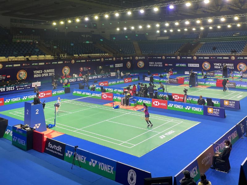 Indoor badminton stadium with multiple courts in use; players compete under bright arena lights and sponsor banners along the sidelines (Yonex Sunrise India Open 2025).