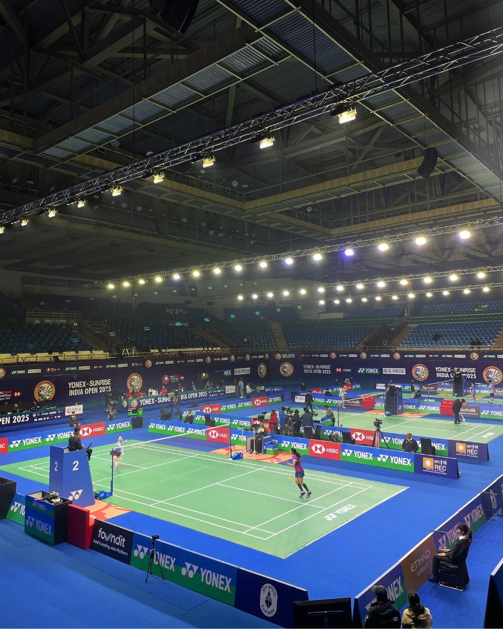 Indoor badminton arena with multiple courts, players in action, and bright overhead lighting announcing Yonex-Sunrise India Open 2025 banners nearby