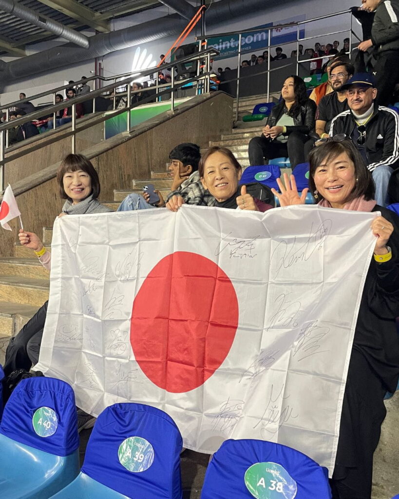 Group of fans in a stadium proudly holding a large signed Japan flag and waving a small flag of Japan.