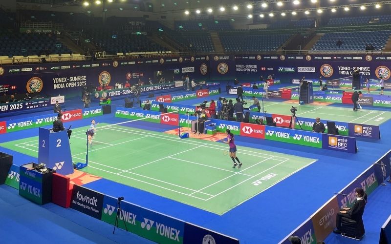 Indoor badminton arena with multiple courts in use for Yonex Sunrise India Open 2025; players compete on green courts while banners line the venue.