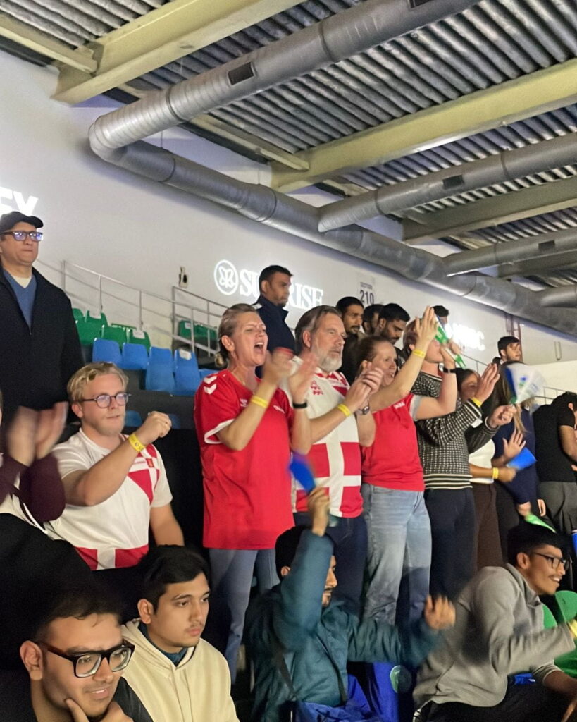 A group of sports fans standing and cheering in a stadium, many wearing red and white jerseys with a Danish flag motif, clapping and raising their hands.