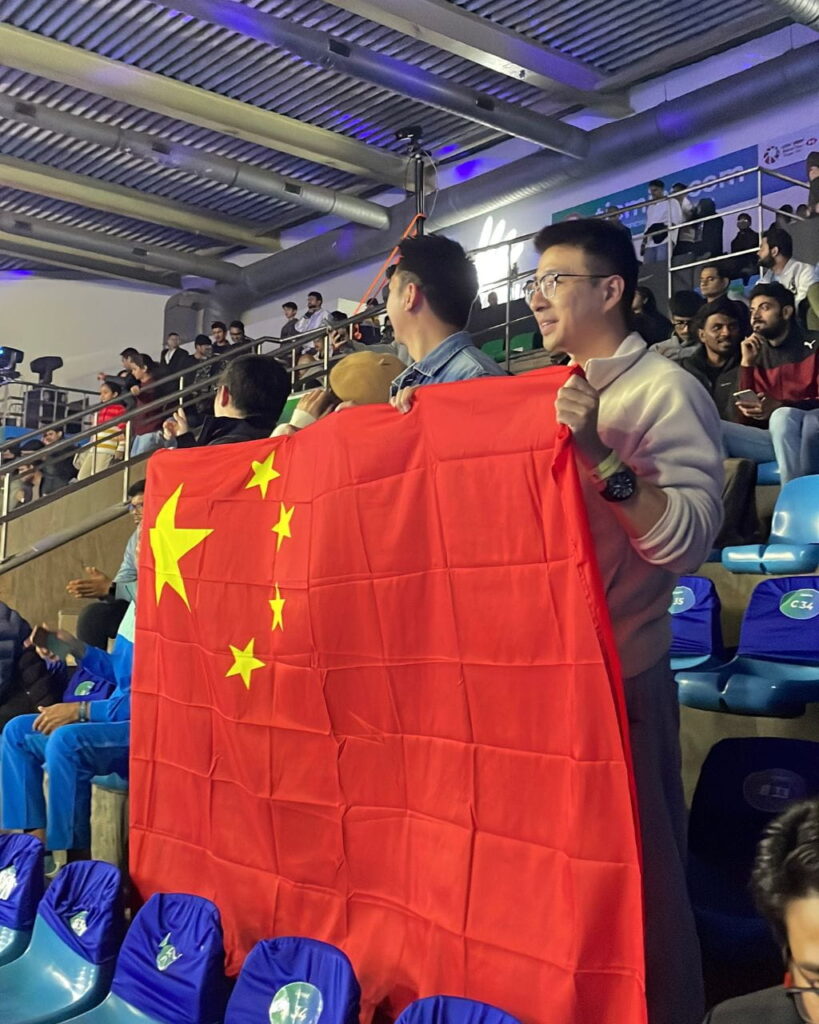 Two spectators waving a large red China flag in a stadium stands area during an event, with others seated around them and arena lighting overhead.