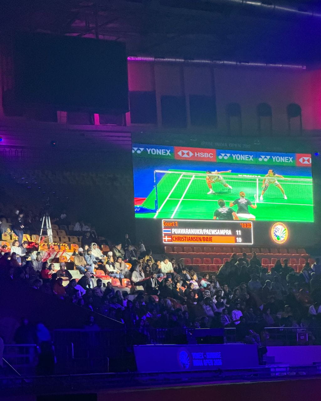 Indoor stadium with a large screen showing a badminton match; players at the net on a bright green court as spectators watch from seats.