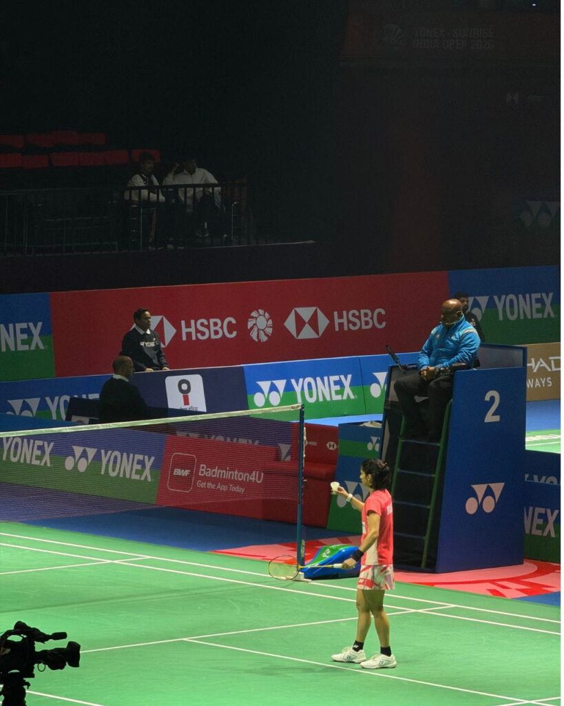 Female badminton player in pink and white stands on the court with racket at the ready; umpire sits on a blue chair nearby amid Yonex banners.