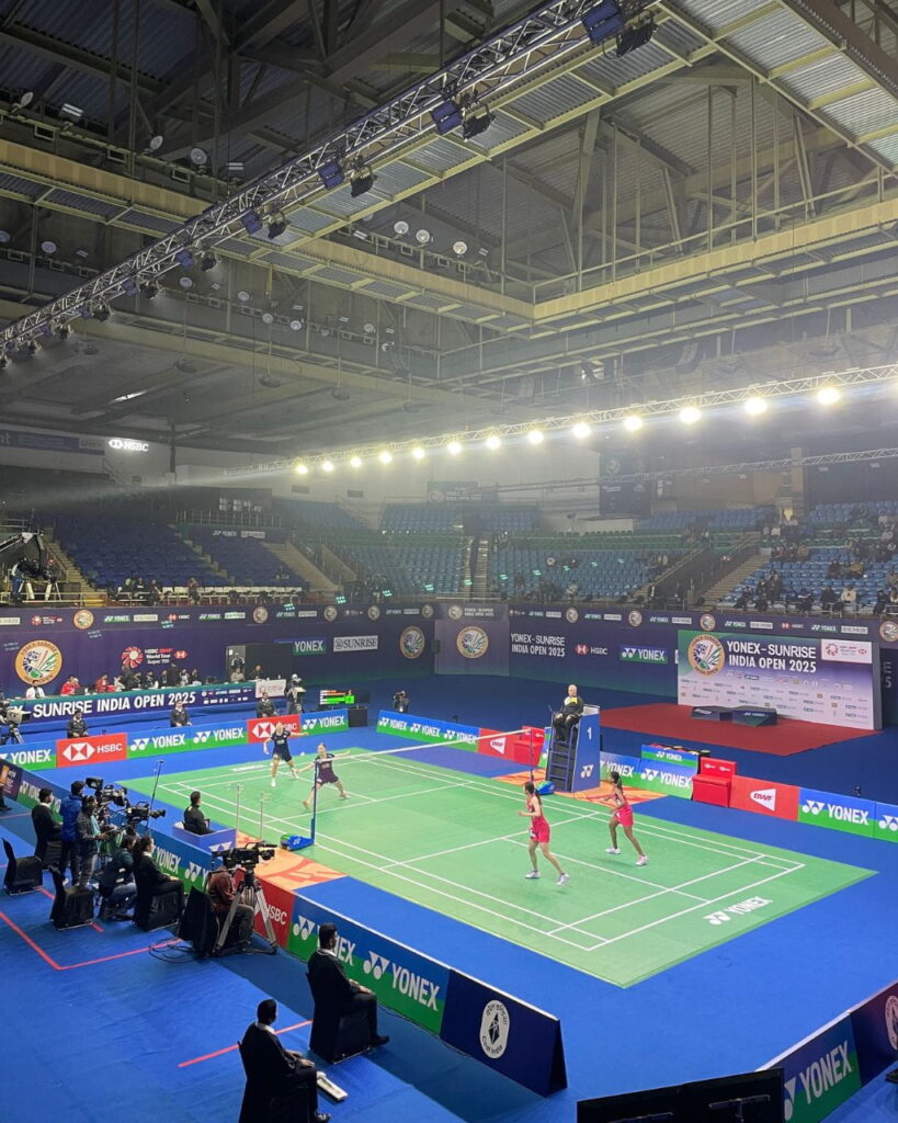 Indoor badminton match at Sunrise India Open 2025; players on a bright blue and green court with officials and cameras around.