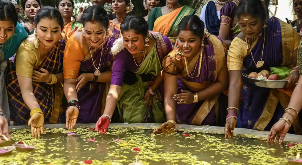 Group of women in colorful sarees perform a traditional ritual around a water tank, scattering flower petals and offerings.