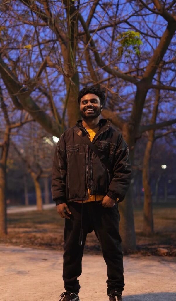 Smiling man in a dark brown jacket with a crossbody bag, standing in a park at dusk among leafless trees.