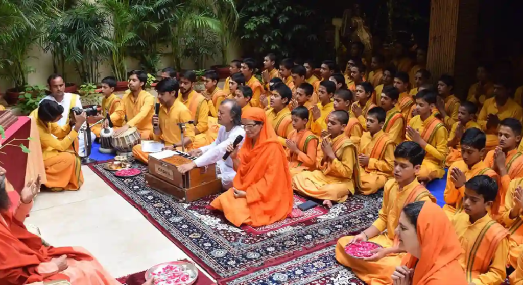 Group of monks in saffron robes seated on a carpet for a religious ceremony; a man with a microphone leads while others pray.