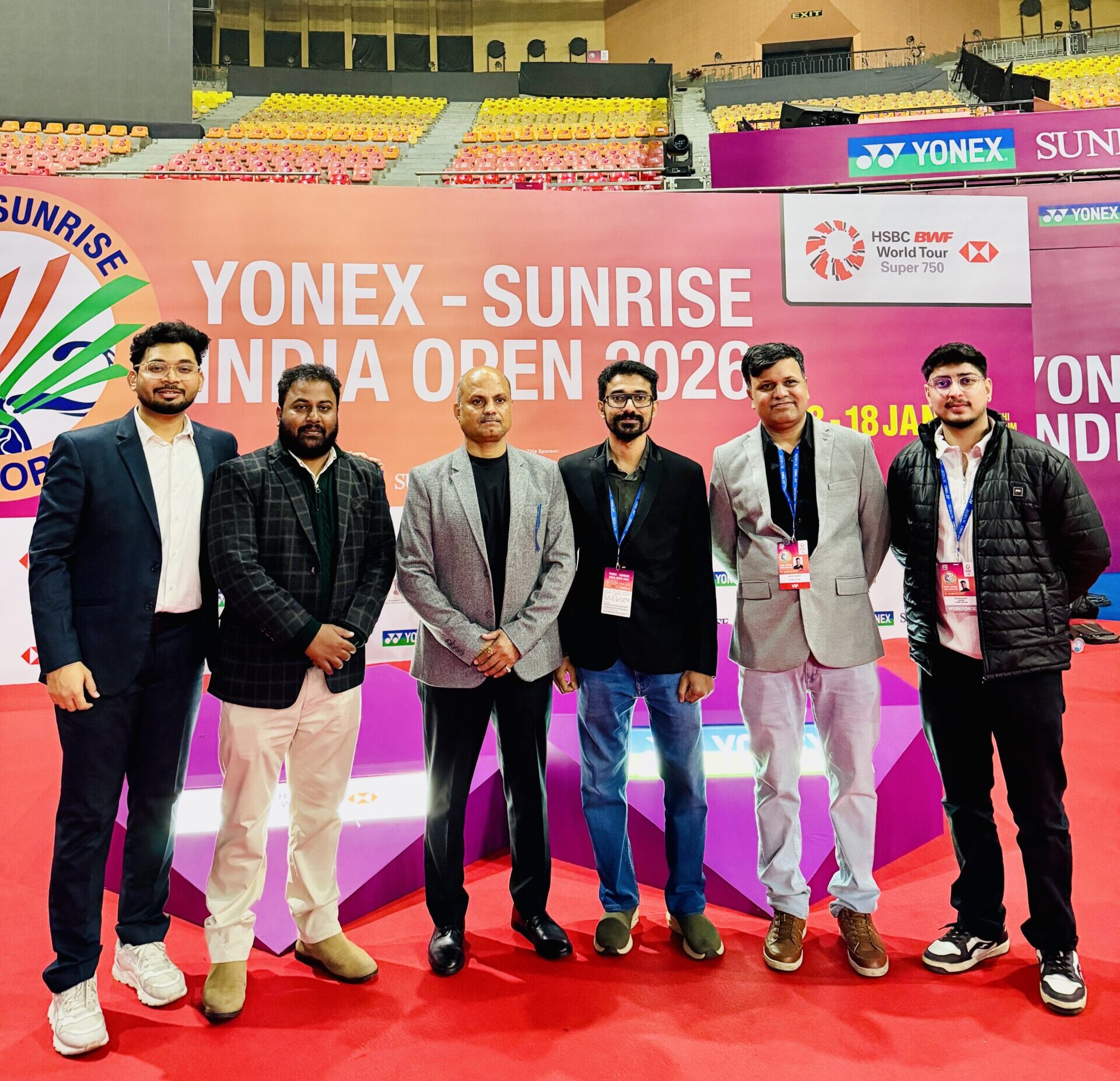 Six men in smart casual attire pose together on a pink stage at a badminton event, with a Yonex-Sunrise India Open 2026 banner in the background.