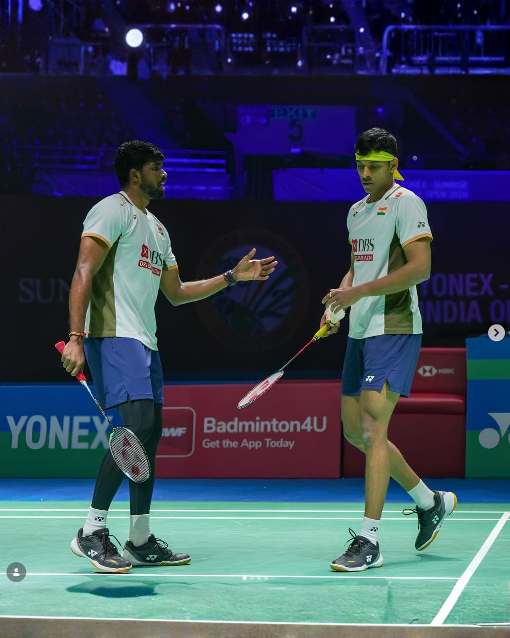 Two Indian badminton players on a court, one gesturing with his racket while the other stands with a headband and shuttle in hand.
