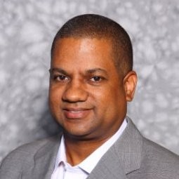 Portrait of a smiling man in a gray suit and white shirt against a neutral patterned backdrop.