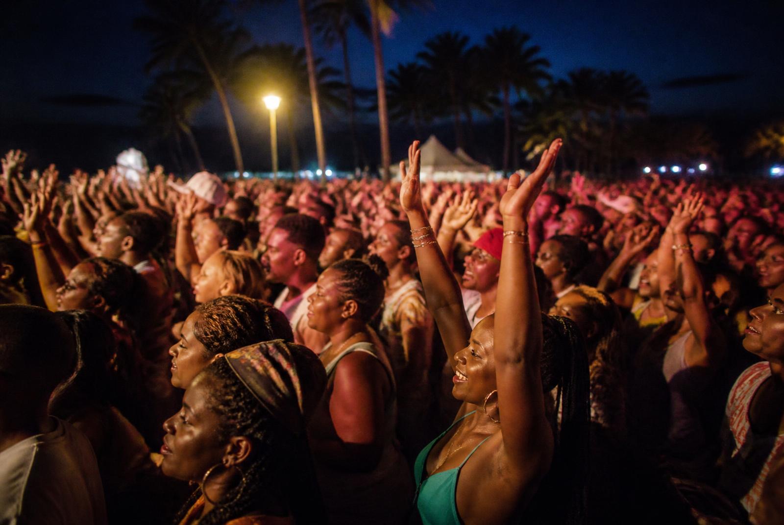 Large crowd at a nighttime outdoor concert, hands raised in the air with palm trees and tents in the background.",