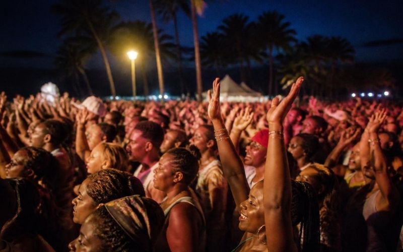 Large crowd at night with many people raising hands, palm trees in background, festival tents visible.