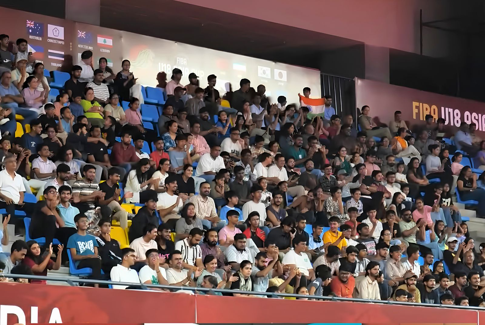 Crowded stadium stands filled with spectators watching a live event; an Indian flag is visible on a spectator’s chair.
